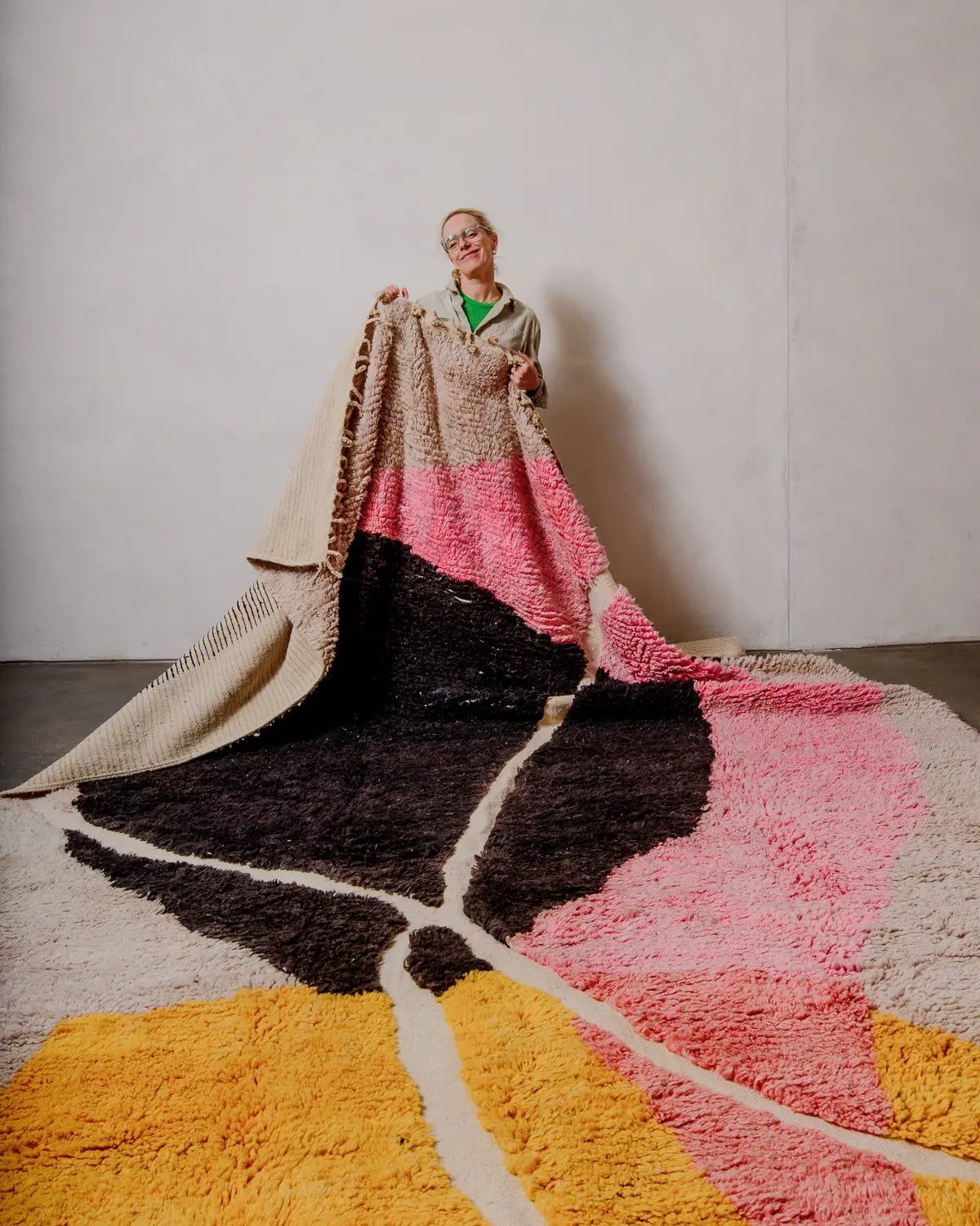 Handcrafted Berberlin Berber rug from Morocco featuring shades of pink, yellow, black and cream being held up against white wall by a woman.